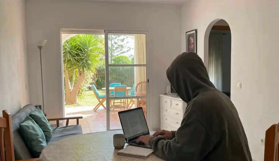 Adam typing on a laptop at a round table in his living room by day, with a sunny garden behind the glass door